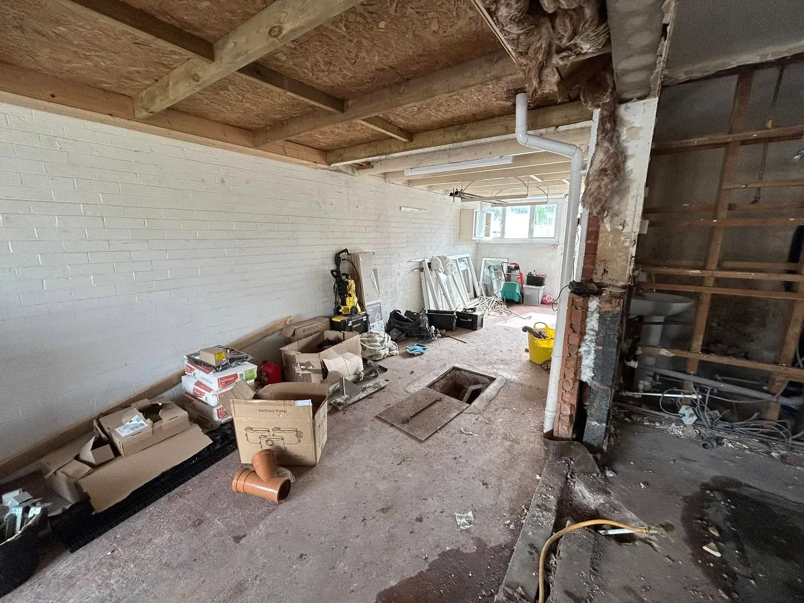 Stripped-out garage interior with exposed roof timbers, an open drainage chamber and boxed materials